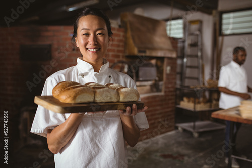 Portrait of smiling asian mid adult female baker holding serving board with fresh breads photo