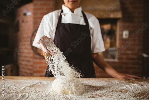 Midsection of asian mid adult female baker sprinkling flour on dough at table in bakery photo