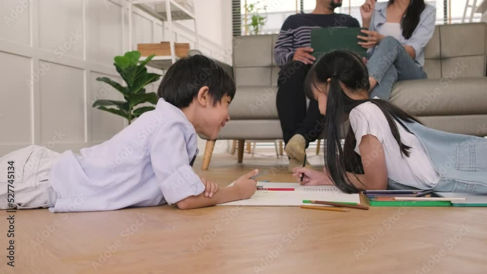 Asian Thai siblings are lying on living room floor, drawing homework with colored pencils together, parents leisurely relax on a sofa, lovely happy weekend activity, and domestic wellbeing lifestyle. Asian Thai siblings are lying on living room floor, drawing homework with colored pencils together, parents leisurely relax on a sofa, lovely happy weekend activity, and domestic wellbeing lifestyle.