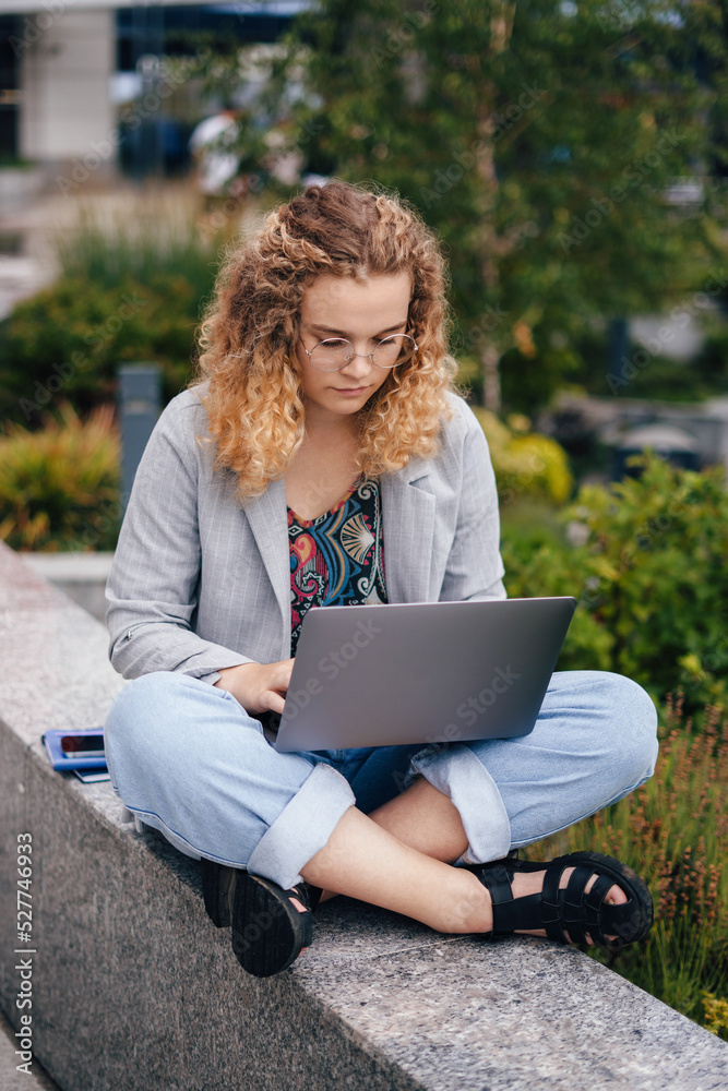 Portrait of a concentrated curly-haired student typing on laptop keyboard, sitting outside the university campus. Education internet technology. Youth lifestyle Portrait of a concentrated curly-haired student typing on laptop keyboard, sitting outside the university campus. Education internet technology. Youth lifestyle