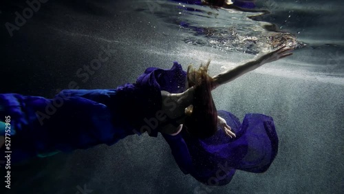 a woman with long hair and wearing a blue dress hovers horizontally and turns around in dark water with bubbles