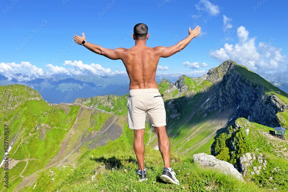 Tanned man with a naked torso in shorts with his arms raised on a mountain top in summer among alpine meadows a tourist on a rock. Travel concept, mountain lovers.