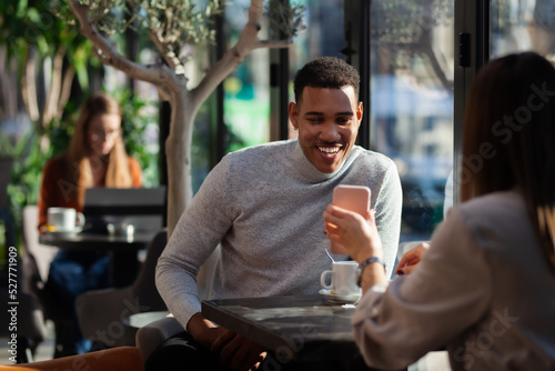 Two friends in a restaurant talking smiling and drinking tea. Multuracial business colleagues having a meeting after work or during coffee break at a cafe bar. Freelance lady working in the background photo