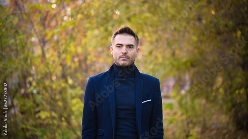 Serious man in a suit stands on the autumn trees in park background.