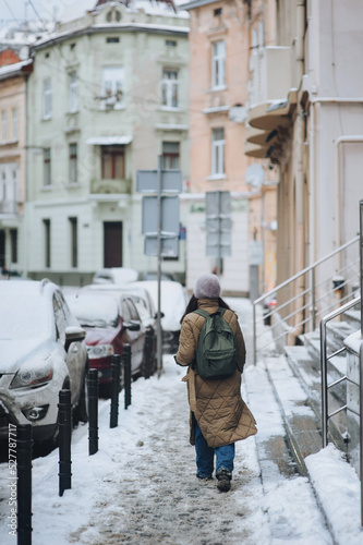 A young Ukrainian woman with a backpack in a down jacket, jeans and an angora hat walks along a snowy winter street with houses and cars. View from the back. Lvov, Ukraine.