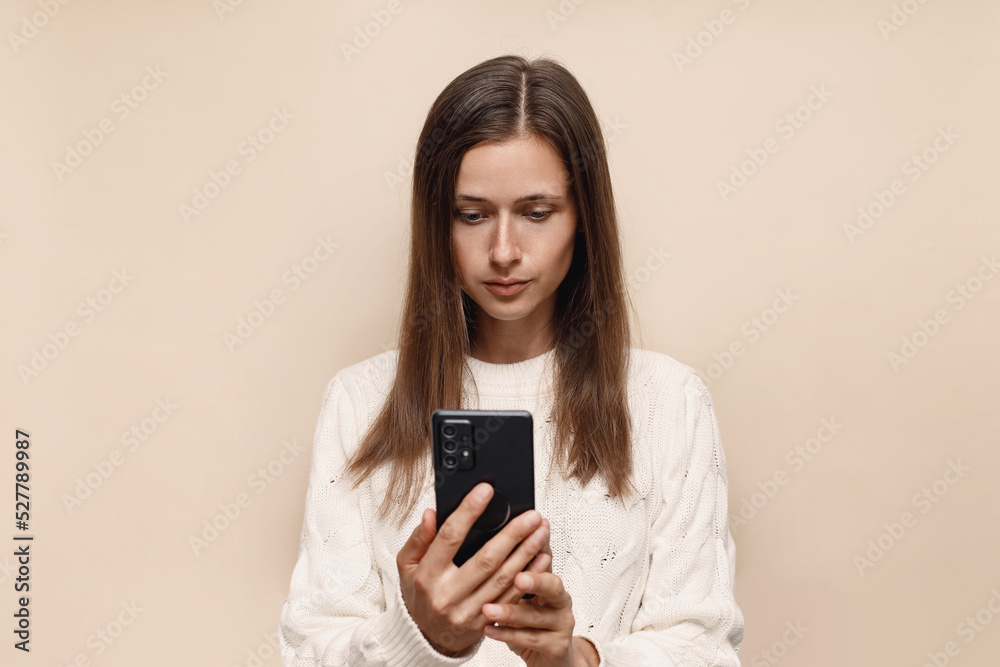 A young caucasian Ukrainian girl looks interestedly into a smartphone, a woman in a white sweater on a beige background A young caucasian Ukrainian girl looks interestedly into a smartphone, a woman in a white sweater on a beige background