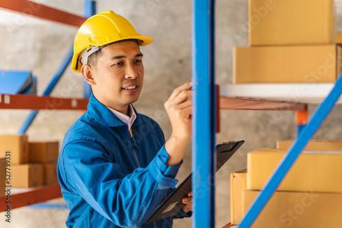 Business Asian warehouse worker checking packages in storehouse and holding paper clipboard.