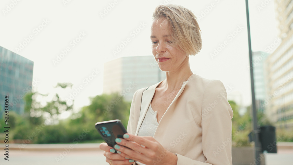 Smiling woman with blond hair wearing beige suit using cellphone while sitting outside on the street Smiling woman with blond hair wearing beige suit using cellphone while sitting outside on the street