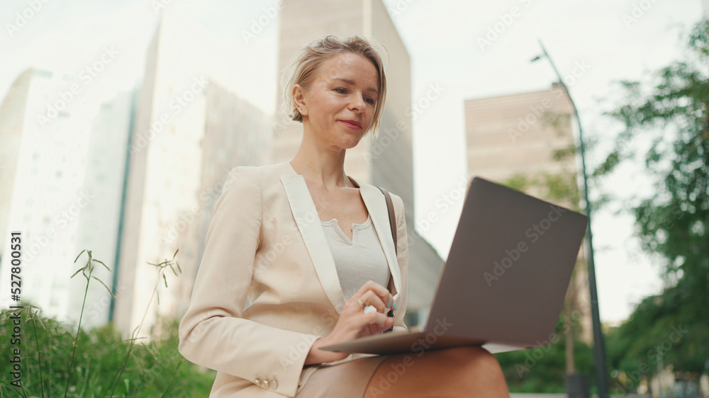 Smiling businesswoman with blond hair wearing beige suit is using pc laptop outside Smiling businesswoman with blond hair wearing beige suit is using pc laptop outside