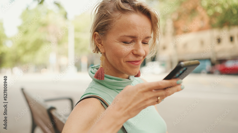 Beautiful happy smiling woman with short blond hair in casual clothes sits on bench, sends voice message on cellphone Beautiful happy smiling woman with short blond hair in casual clothes sits on bench, sends voice message on cellphone