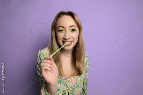 Fashionable young woman with bright makeup chewing bubblegum on lilac background