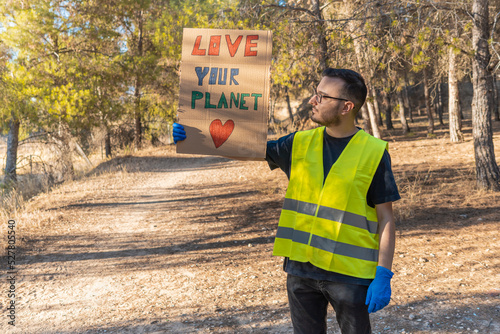 Hombre voluntario, con pancarta de ama tu planeta, en el bosque, en forma de protesta. Fotografía horizontal con espacio para texto.