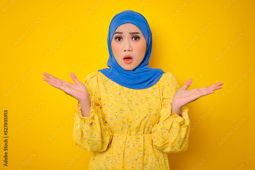 Shocked young Asian woman in casual dress looking at camera with opened mouth, spreading hands isolated on yellow background Shocked young Asian woman in casual dress looking at camera with opened mouth, spreading hands isolated on yellow background