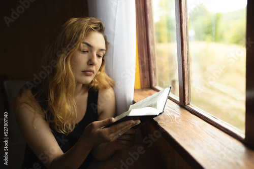 The girl sits by the window and reads a book. Reading. The girl is reading an old book. Time for yourself. Relax and rest while reading.