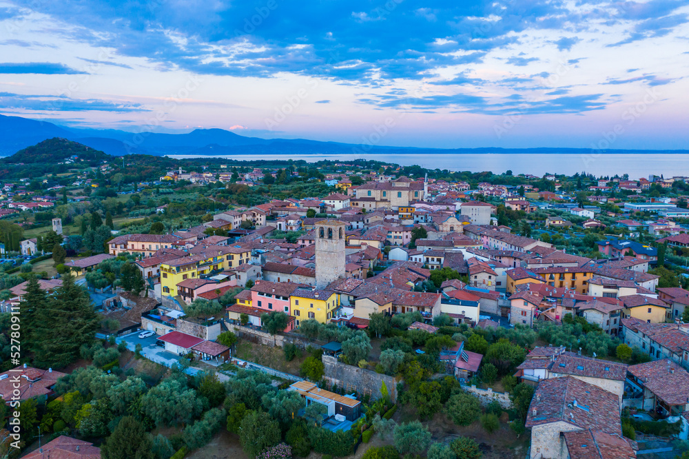 Obraz premium Aerial view of village near Lago di Garda Italy. Beautiful sunset light. Flying close to the church tower revealing Lago di Garda on the horizon. Manerba del Garda town in Brescia Italy. 