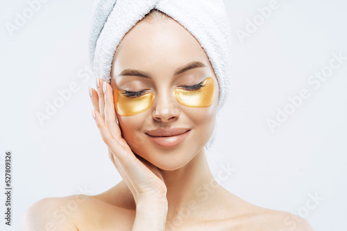 Close up portrait of charming young woman with cosmetic collagen patches under eyes  enjoys flawless of skin  has well cared complexion  manicure  wears wrapped towel on head  stands indoor.
