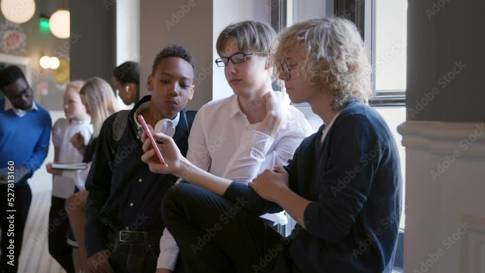 Multiracial young friends using mobile phone while sitting on windowsill in school corridor Multiracial young friends using mobile phone while sitting on windowsill in school corridor