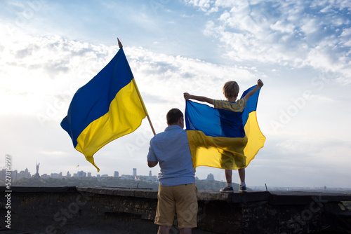 dad and son stand together on the high roof of a house in the city of Kyiv with two flags of Ukraine against the sky