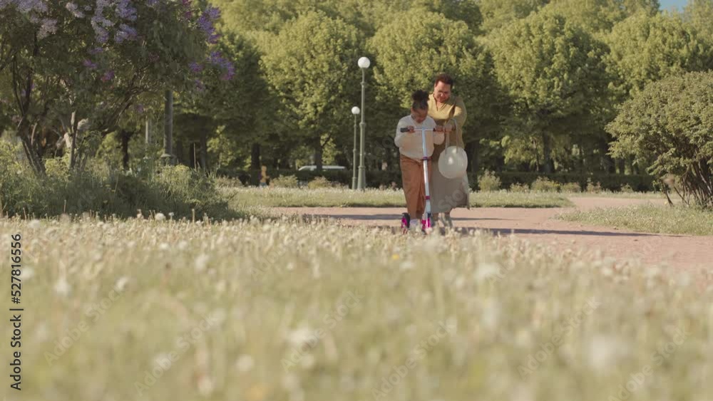 Full length slowmo of African American mother teaching her little daughter to ride scooter along pathway in park on warm sunny day Full length slowmo of African American mother teaching her little daughter to ride scooter along pathway in park on warm sunny day