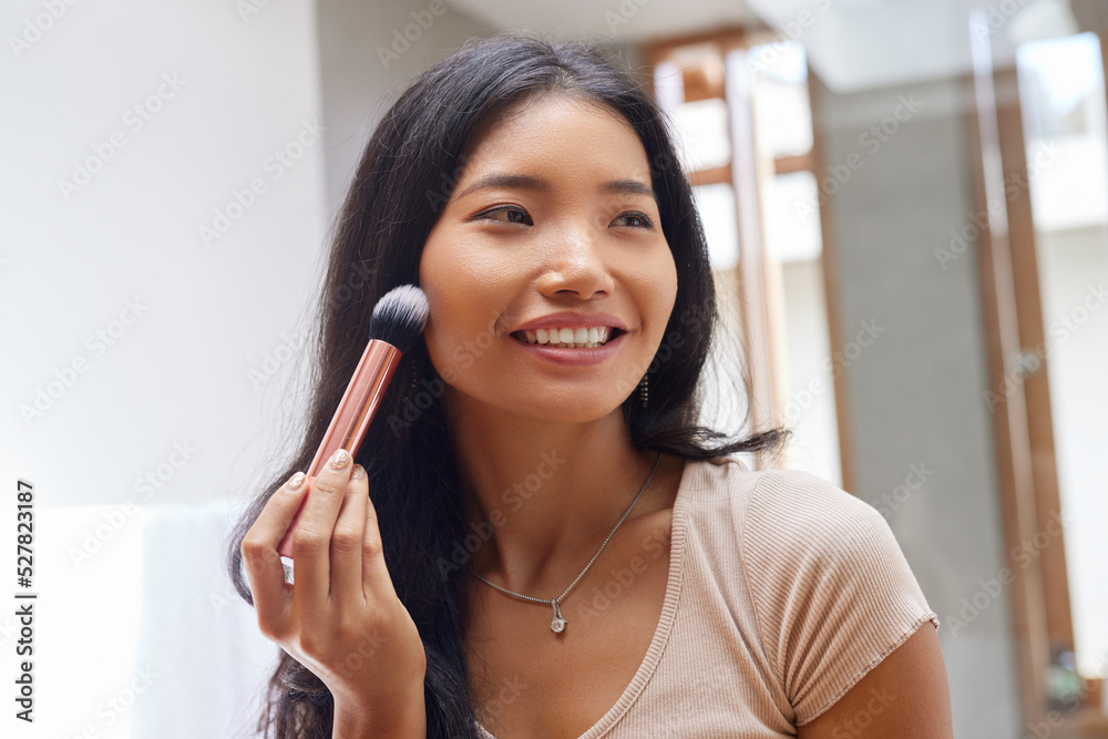 Beauty Makeup. Smiling Woman Applying Cosmetics on Face with Brush in Front of Mirror at Bathroom. Portrait of Happy Asian Girl Model Doing Make up in Morning 