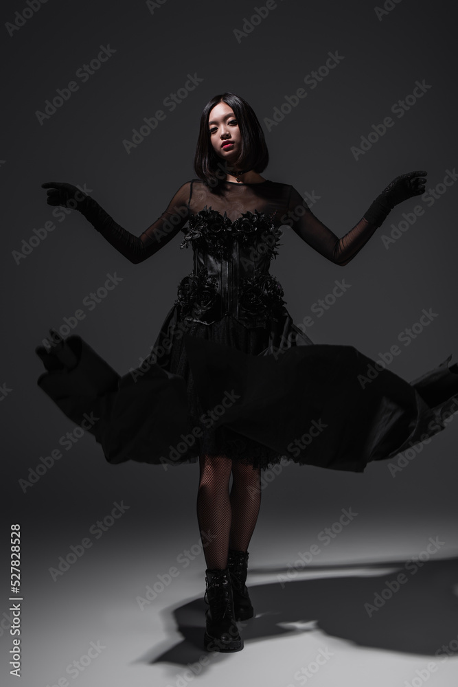 full length of asian young woman in black gothic dress and gloves gesturing on dark grey.