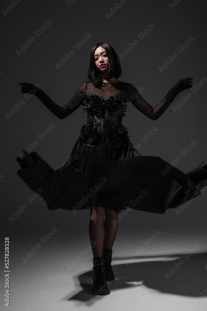 full length of asian young woman in black gothic dress and gloves gesturing on dark grey. full length of asian young woman in black gothic dress and gloves gesturing on dark grey.