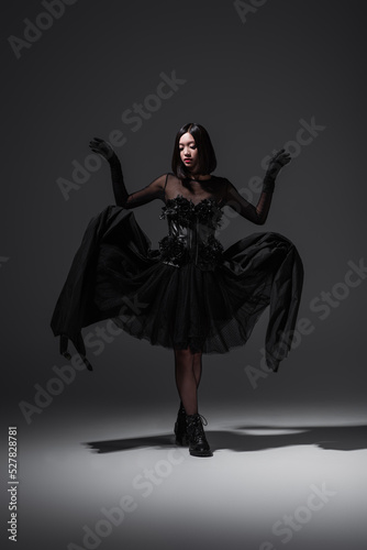 full length of asian young woman in black gothic dress and gloves posing on dark grey.