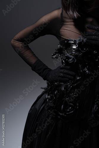 cropped view of woman in black gothic dress and gloves posing with hand on hip on dark grey.
