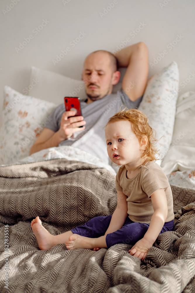 Young man father laying on the bed using cellphone with his toddler child sitting on foreground and looking disappointed. Kid feeling lonely. Selective focus.