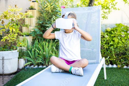 Little girl wearing virtual reality glasses, holding them with her hands, looking straight ahead, sitting on a deckchair in the garden of her house. Metaverse, VR, game, digital, simulation concept.