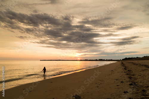 Jeune fille ou femme se promenant au bord de la mer, les pieds dans l'eau, vers le soleil couchant, à contre-jour.