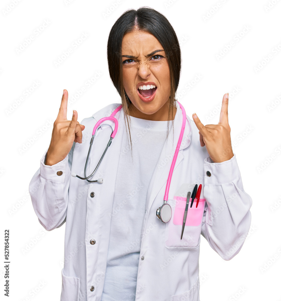 Beautiful hispanic woman wearing doctor uniform and stethoscope shouting with crazy expression doing rock symbol with hands up. music star. heavy concept. Beautiful hispanic woman wearing doctor uniform and stethoscope shouting with crazy expression doing rock symbol with hands up. music star. heavy concept.