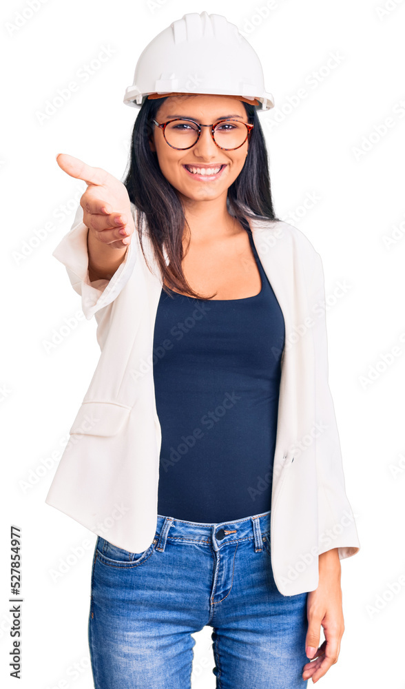 Young beautiful latin girl wearing architect hardhat and glasses smiling friendly offering handshake as greeting and welcoming. successful business. Young beautiful latin girl wearing architect hardhat and glasses smiling friendly offering handshake as greeting and welcoming. successful business.