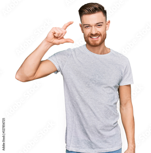 Young redhead man wearing casual grey t shirt smiling and confident gesturing with hand doing small size sign with fingers looking and the camera. measure concept.