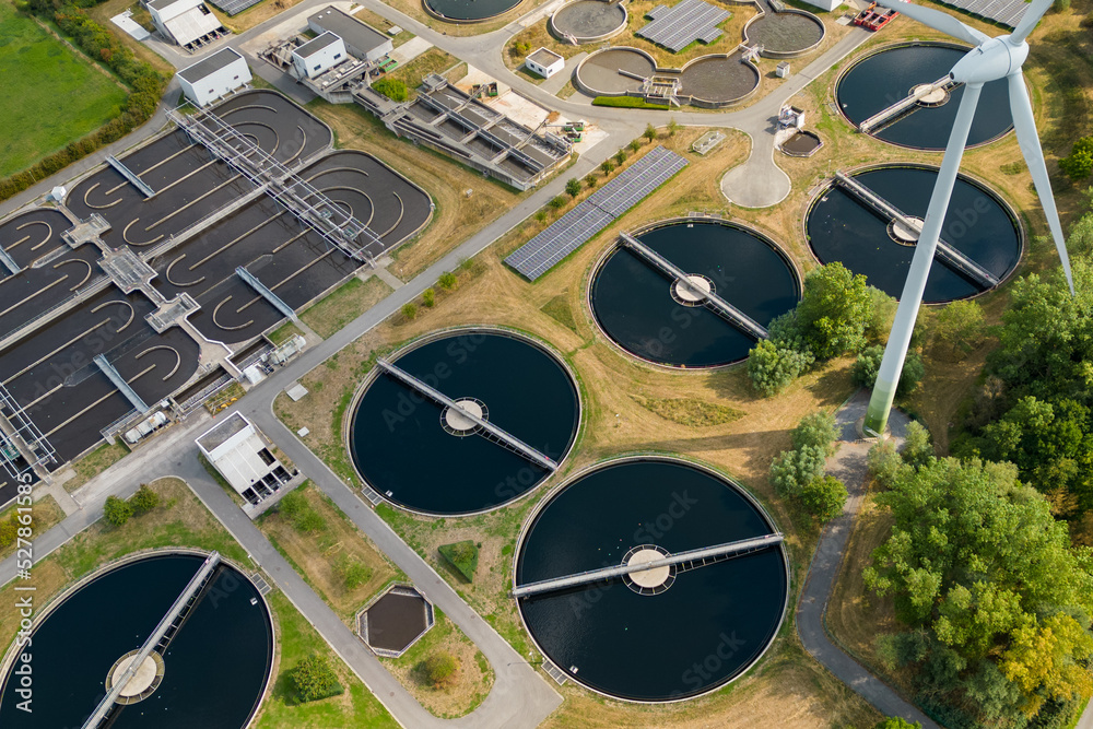 Aerial view of storage tanks in a modern sewage water treatment plant to recycle waste water. Wind turbine present on the industrial plant. Aerial view of storage tanks in a modern sewage water treatment plant to recycle waste water. Wind turbine present on the industrial plant.