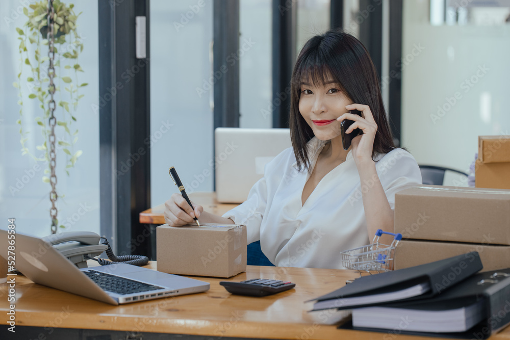 Asian businesswoman using smartphone with parcel boxes and checking orders sitting at office desk. Business online selling and online shopping concepts. Asian businesswoman using smartphone with parcel boxes and checking orders sitting at office desk. Business online selling and online shopping concepts.