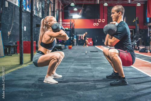 Slika na platnu Fit couple in squat position standing face to face lifting a kettle bells during exercise class in the gym