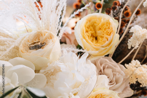 Rustic-style wedding rings lie in a white peony bud. Nearby are cereal grasses, spikelets, yellow peonies, and roses.