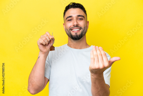Canvas Print Young Arab man holding home keys isolated on yellow background inviting to come with hand