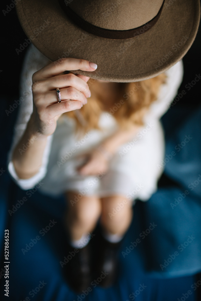 A girl in a white dress sits on a blue sofa. In the foreground is a brown hat, the girl holds the brim of the hat with her hand.