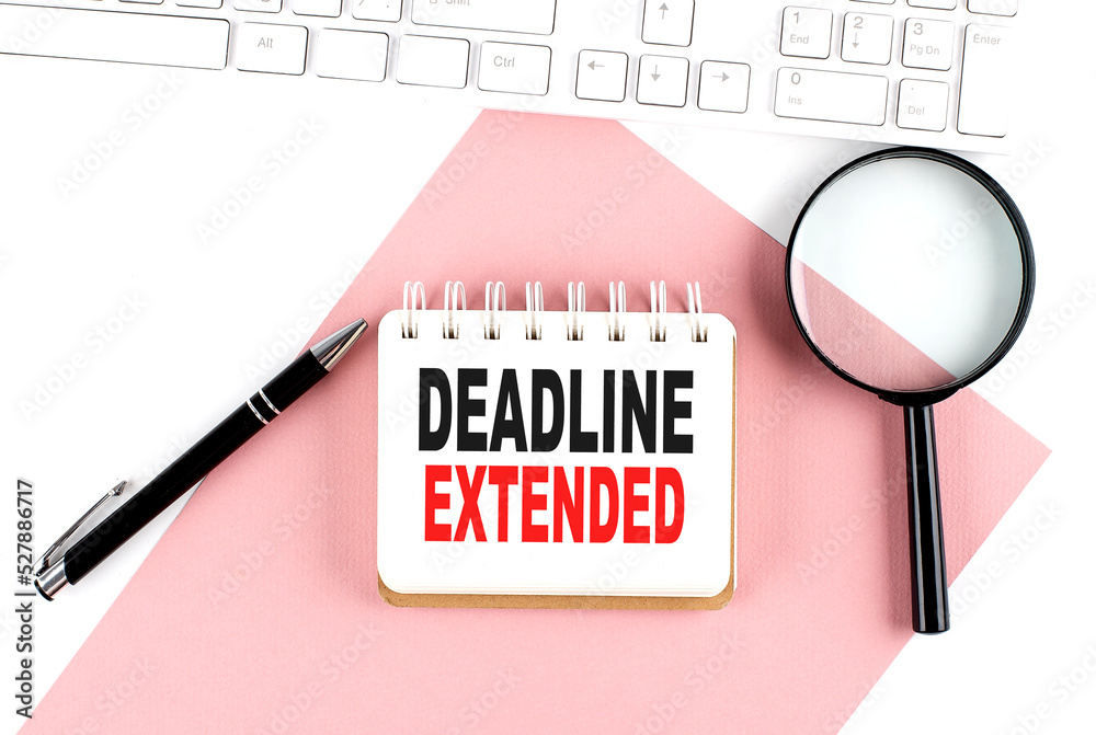 Business concept. Notebook with text DEADLINE EXTENDED on pink paper with magnifier, keyboard and pen in the white background Business concept. Notebook with text DEADLINE EXTENDED on pink paper with magnifier, keyboard and pen in the white background