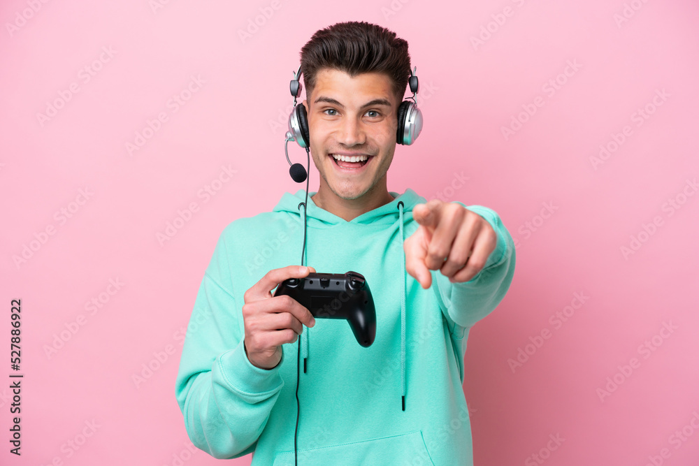 Young handsome caucasian man playing with a video game controller isolated on pink background surprised and pointing front Young handsome caucasian man playing with a video game controller isolated on pink background surprised and pointing front