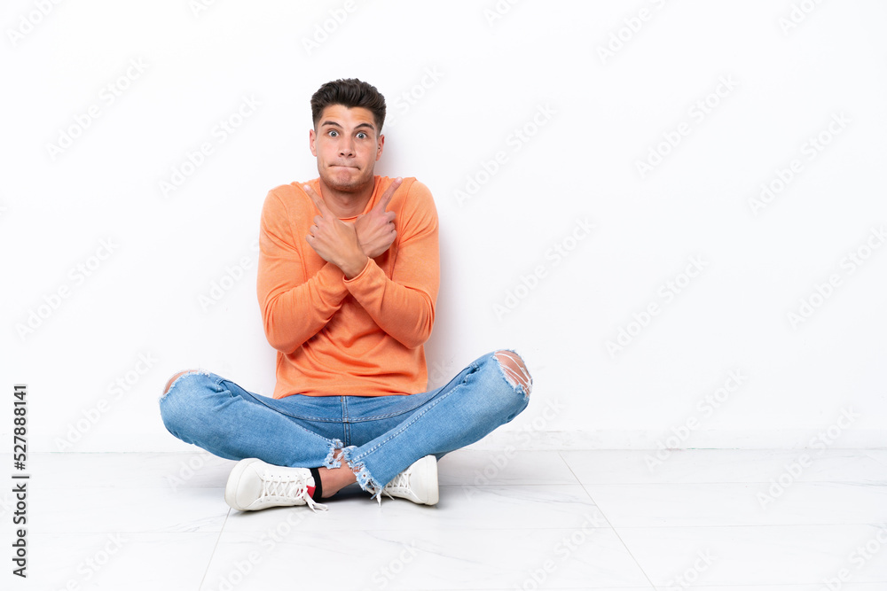 Young man sitting on the floor isolated on white background pointing to the laterals having doubts