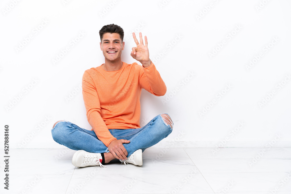 Young man sitting on the floor isolated on white background happy and counting three with fingers