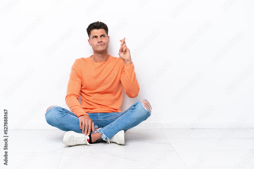 Young man sitting on the floor isolated on white background with fingers crossing and wishing the best Young man sitting on the floor isolated on white background with fingers crossing and wishing the best
