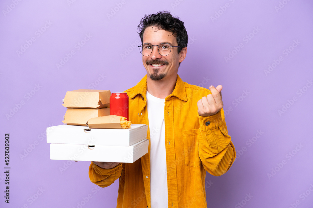 Young caucasian man holding fast food isolated on purple background making money gesture Young caucasian man holding fast food isolated on purple background making money gesture
