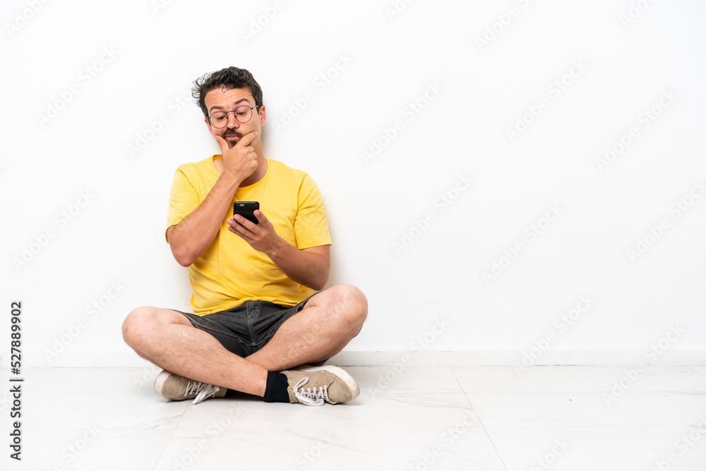 Fototapeta premium Young caucasian man sitting on the floor isolated on white background thinking and sending a message