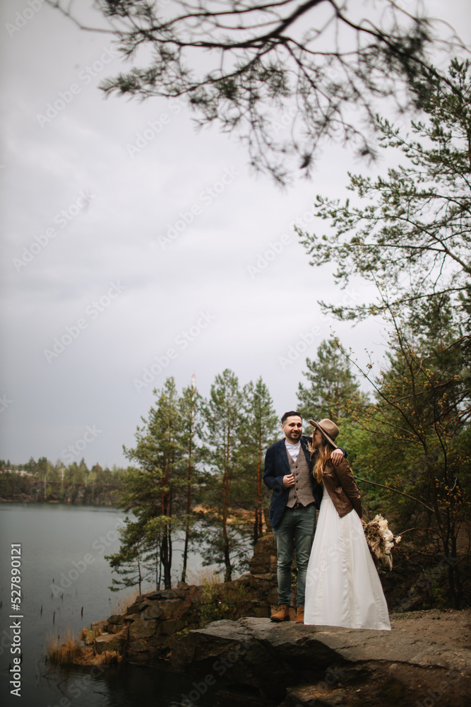 The newlyweds stand on a canyon rock in the forest near the lake. The bride in a white dress, leather jacket and hat, the groom hugs her. The bride and groom laugh in nature. The newlyweds stand on a canyon rock in the forest near the lake. The bride in a white dress, leather jacket and hat, the groom hugs her. The bride and groom laugh in nature.