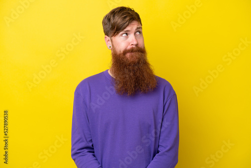 Redhead man with beard isolated on yellow background having doubts while looking up