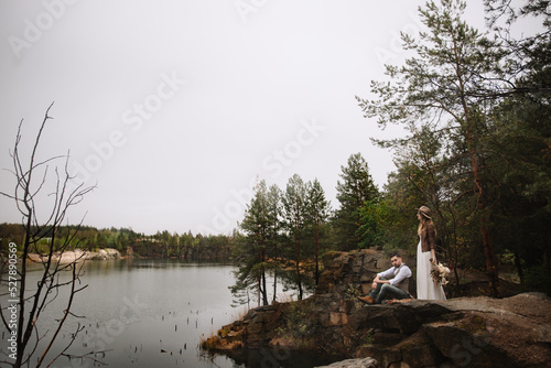 The bride and groom are sitting in a stone canyon by the lake. The groom is sitting on a rock. The bride in a white dress, leather jacket, and hat holds a wedding bouquet and looks at the lake.